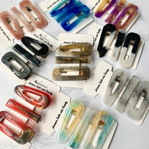May include: A collection of resin hair clips in various colors, including pink, blue, purple, black, and gold. The clips are rectangular and feature gold-colored metal clasps. Each clip is displayed on a small card with the text "Amreva Bath and Beauty".