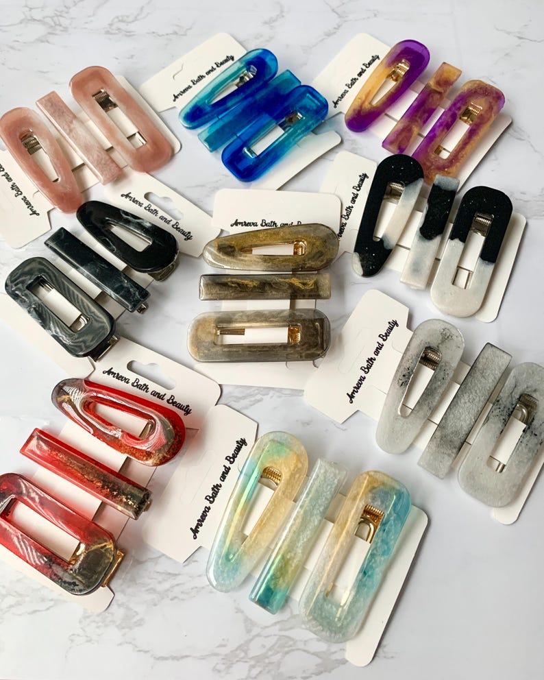 May include: A collection of resin hair clips in various colors, including pink, blue, purple, black, and gold. The clips are rectangular and feature gold-colored metal clasps. Each clip is displayed on a small card with the text "Amreva Bath and Beauty".