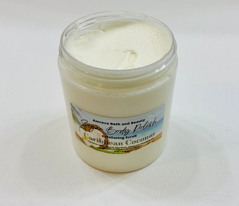 May include: A white jar of sugar body polish with a label that reads "Sugar Body Polish Exfoliating Scrub Caribbean Coconut" by Amreva Bath and Beauty.