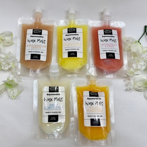 May include: Five squeeze pouches of wax melts in various colors, including tan, yellow, and orange. Each pouch has a label with the scent name: Gingerbread Cookie, Sunshine, Grapefruit Citrus, Ode 2 Eliminator, and Peach Passion. The pouches are arranged on a white surface with white flowers.