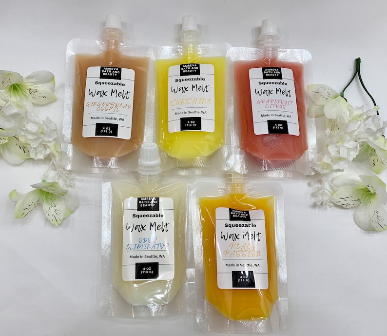May include: Five squeeze pouches of wax melts in various colors, including tan, yellow, and orange. Each pouch has a label with the scent name: Gingerbread Cookie, Sunshine, Grapefruit Citrus, Ode 2 Eliminator, and Peach Passion. The pouches are arranged on a white surface with white flowers.