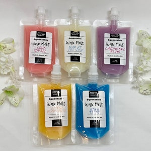 May include: An assortment of five squeeze pouches of scented wax melts in various colors, including pink, white, purple, yellow, and blue. Each pouch has a white label with the product name and brand. The pouches are arranged on a white surface with white flowers.