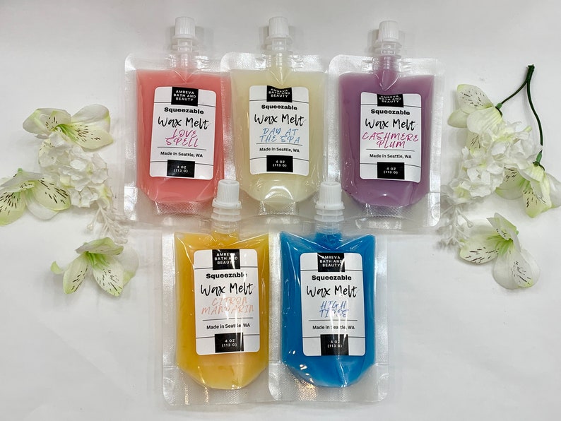 May include: An assortment of five squeeze pouches of scented wax melts in various colors, including pink, white, purple, yellow, and blue. Each pouch has a white label with the product name and brand. The pouches are arranged on a white surface with white flowers.