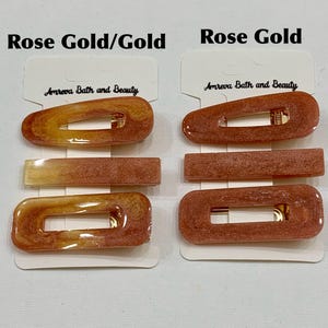May include: Hair clips in rose gold and gold tones. The clips are rectangular and oval-shaped, with a gold-colored clasp. The brand name "Amreva Bath and Beauty" is printed on the packaging.