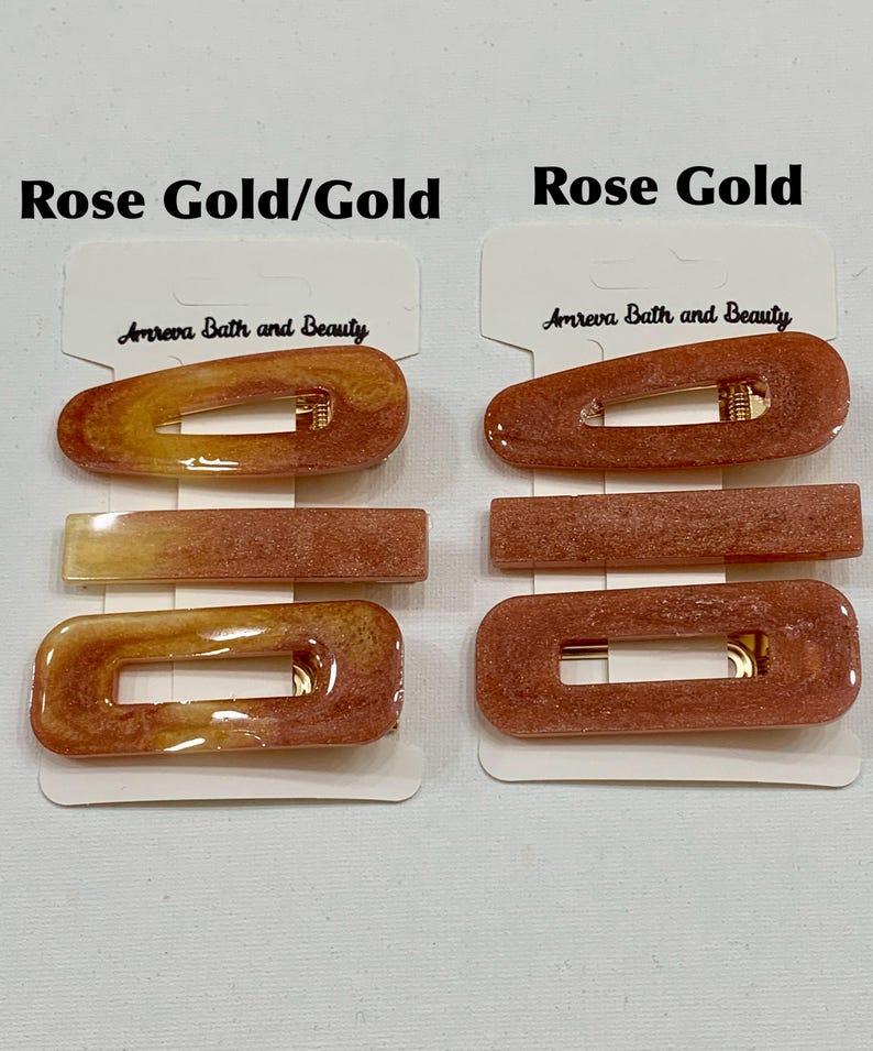May include: Hair clips in rose gold and gold tones. The clips are rectangular and oval-shaped, with a gold-colored clasp. The brand name "Amreva Bath and Beauty" is printed on the packaging.