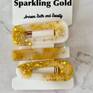 May include: Three gold and white hair clips with gold glitter accents. The clips are rectangular and oval-shaped, with a gold metal clasp. The top of the packaging reads "Sparkling Gold" and "Amreva Bath and Beauty".