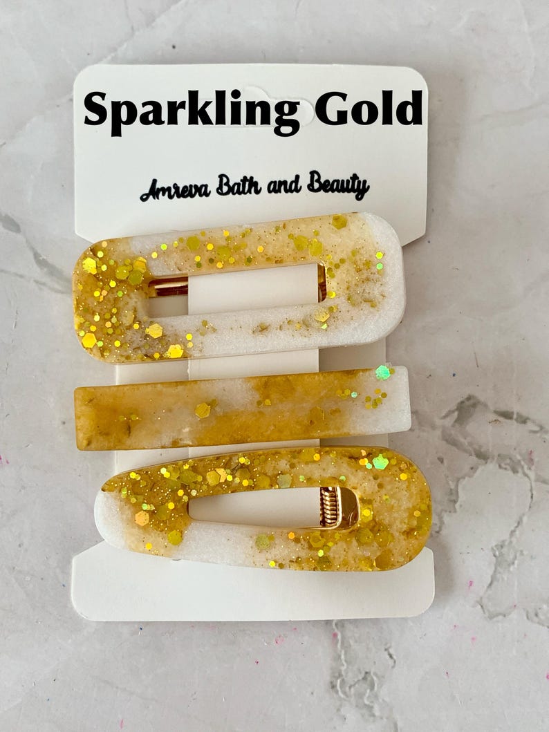 May include: Three gold and white hair clips with gold glitter accents. The clips are rectangular and oval-shaped, with a gold metal clasp. The top of the packaging reads "Sparkling Gold" and "Amreva Bath and Beauty".