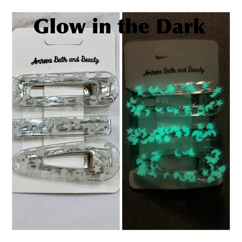 May include: Three clear hair clips with silver metal clasps, filled with moon and star shapes. The clips are on a white card with the text "Glow in the Dark" and "Amreva Bath and Beauty". The right side of the image shows the clips glowing green.