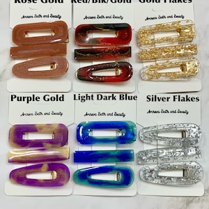 May include: A collection of hair clips in various colors and styles. The clips include rose gold, red/black/gold, gold flakes, purple gold, light dark blue, and silver flakes. Each clip is labeled with the "Amreva Bath and Beauty" brand.