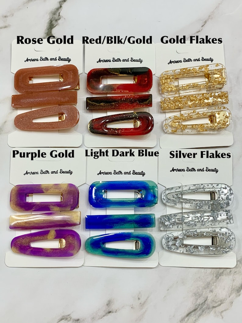 May include: A collection of hair clips in various colors and styles. The clips include rose gold, red/black/gold, gold flakes, purple gold, light dark blue, and silver flakes. Each clip is labeled with the "Amreva Bath and Beauty" brand.
