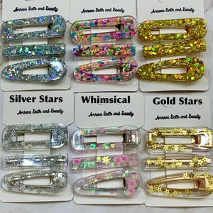 May include: A collection of hair clips in various designs, including silver sequin, sprinkles, gold sequin, silver stars, whimsical, and gold stars. Each clip is rectangular and features decorative elements. The brand name "Amreva Bath and Beauty" is printed on the tags.