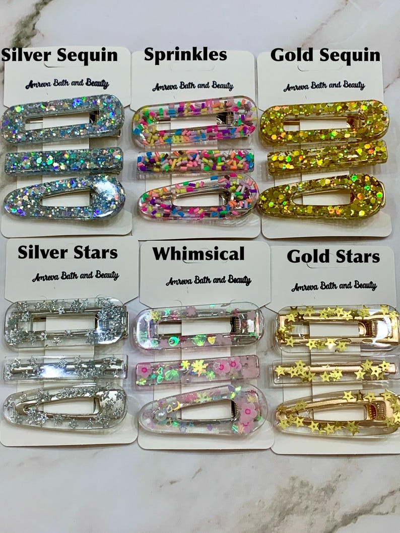 May include: A collection of hair clips in various designs, including silver sequin, sprinkles, gold sequin, silver stars, whimsical, and gold stars. Each clip is rectangular and features decorative elements. The brand name "Amreva Bath and Beauty" is printed on the tags.
