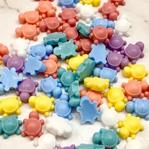 May include: A collection of small, colorful turtle-shaped objects in various shades of pink, red, blue, green, yellow, purple, and white. The turtles are scattered across a white surface, creating a playful and vibrant display.