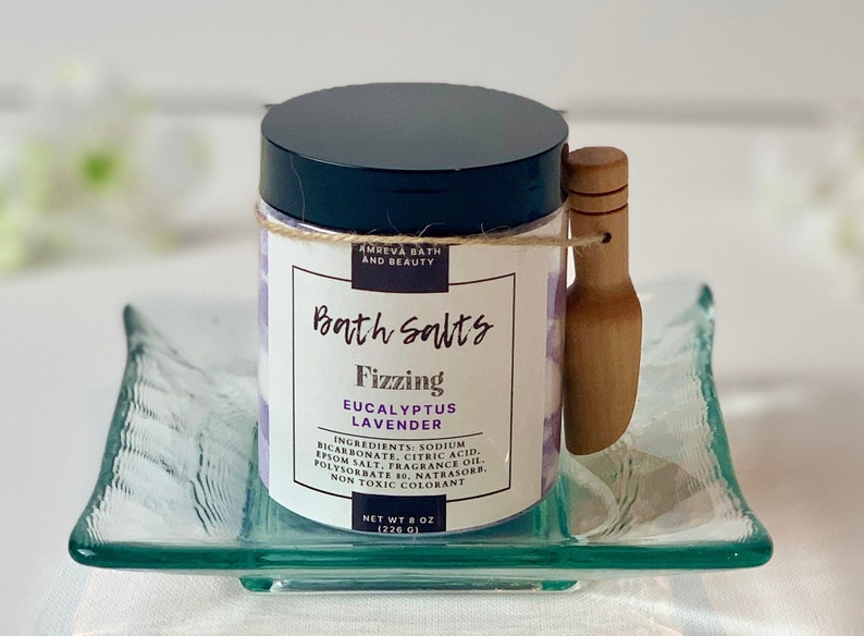 Pode incluir: Um frasco de sais de banho com uma concha de madeira. O frasco &eacute; rotulado como "Bath Salts Fizzing Eucalyptus Lavender" e lista os ingredientes. O frasco est&aacute; sobre um prato de vidro transparente.