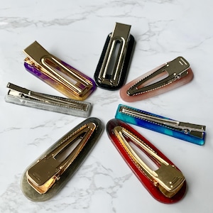 May include: A collection of seven hair barrettes in various colors, including purple, black, pink, blue, red, silver, and gray. Each barrette features a gold-colored metal clasp. The barrettes are arranged in a circular pattern.