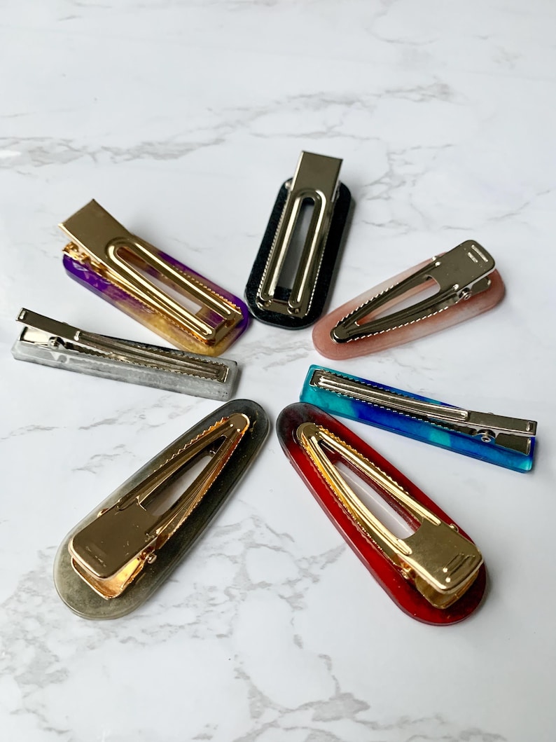 May include: A collection of seven hair barrettes in various colors, including purple, black, pink, blue, red, silver, and gray. Each barrette features a gold-colored metal clasp. The barrettes are arranged in a circular pattern.