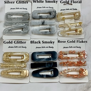 May include: A collection of hair clips in various styles and colors, including silver glitter, white smoky, gold floral, gold glitter, black smoky, and rose gold flakes. Each clip is displayed on a white card with the text "Amreva Bath and Beauty".