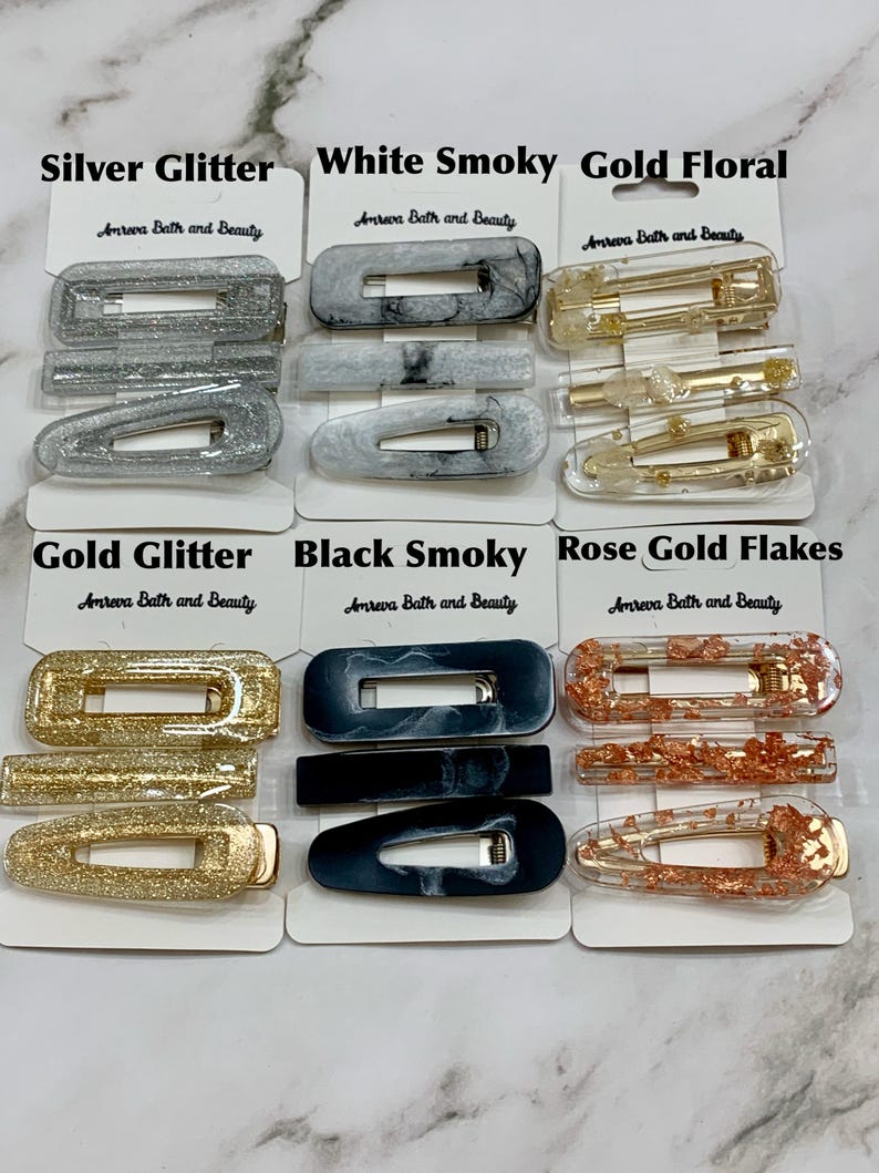 May include: A collection of hair clips in various styles and colors, including silver glitter, white smoky, gold floral, gold glitter, black smoky, and rose gold flakes. Each clip is displayed on a white card with the text "Amreva Bath and Beauty".