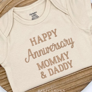 May include: A beige baby onesie with the text "HAPPY Anniversary MOMMY & DADDY" in brown lettering.