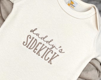 May include: A cream-colored baby onesie with the text "Daddy's Sidekick" embroidered in brown.