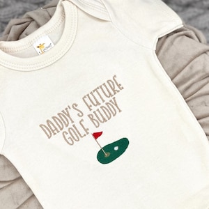 May include: A white baby onesie with the text "Daddy's Future Golf Buddy" in brown lettering. A green golf course with a red flag is embroidered on the onesie.