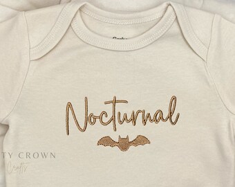 May include: A cream-colored baby onesie with the word "Nocturnal" embroidered in brown script with a brown bat underneath.