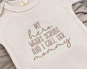 May include: A cream-colored baby onesie with the text "My hero wears scrubs and I call her mommy" embroidered in brown.