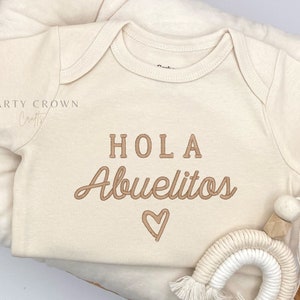 May include: A beige baby onesie with the words "HOLA Abuelitos" embroidered in brown, along with a heart symbol. The onesie is placed on a soft, cream-colored cushion, with a small rainbow-shaped decoration.