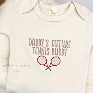 May include: A cream-colored baby onesie with the text "Daddy's Future Tennis Buddy" embroidered in brown. Two red tennis rackets are embroidered below the text.
