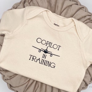 May include: A cream-colored baby onesie with black embroidery that reads "Copilot in Training" and a small airplane graphic.
