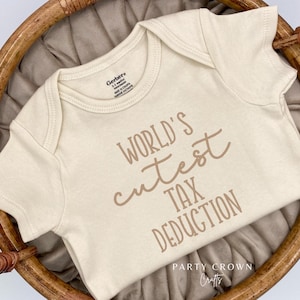 May include: A white onesie with the text "World's Cutest Tax Deduction" in brown lettering. The onesie is in a wicker basket.