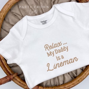 May include: White baby onesie with brown text that reads "Relax... My Daddy is a Lineman".