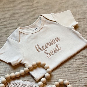 Puede incluir: Un body blanco para beb&eacute; con las palabras "Heaven Sent" bordadas en hilo marr&oacute;n. El body est&aacute; sobre una tela beige texturizada con una guirnalda de cuentas de madera.