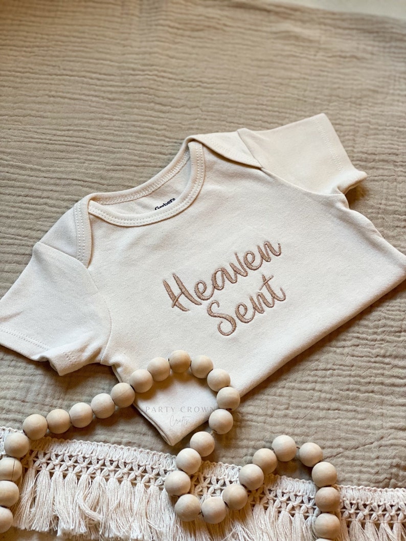 Puede incluir: Un body blanco para beb&eacute; con las palabras "Heaven Sent" bordadas en hilo marr&oacute;n. El body est&aacute; sobre una tela beige texturizada con una guirnalda de cuentas de madera.