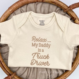 May include: A cream-colored baby onesie with the text "Relax... My Daddy is a Truck Driver" printed in brown. The onesie is laid out on a woven basket.