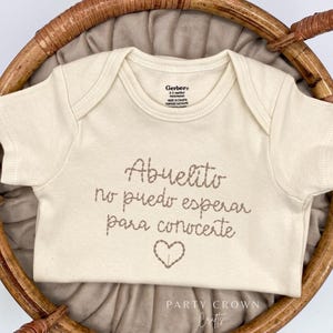 May include: A white onesie with the text "Abuelito no puedo esperar para conocerte" in brown stitching. A brown heart is stitched below the text. The onesie is lying on a brown wicker basket.
