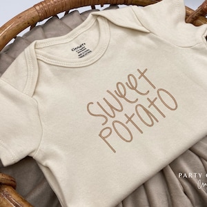 May include: A cream-colored baby onesie with short sleeves and the text "Sweet Potato" in brown lettering.
