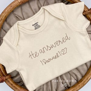 K&ouml;nnte beinhalten: Cremefarbener Baby-Body mit der gestickten Aufschrift "He answered 1 Samuel 1:27." Der Body besteht aus weichem Material und wird in einem Weidenkorb pr&auml;sentiert. Das Markenetikett "Gerber" ist sichtbar.