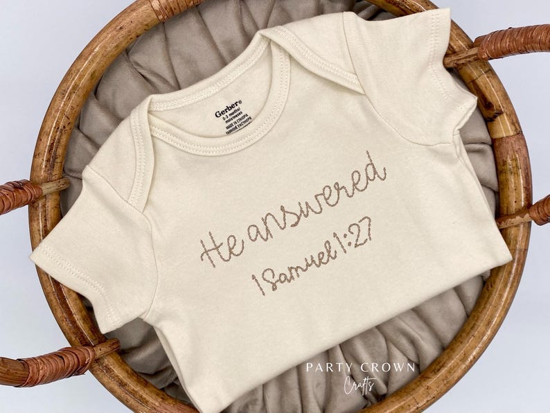 K&ouml;nnte beinhalten: Cremefarbener Baby-Body mit der gestickten Aufschrift "He answered 1 Samuel 1:27." Der Body besteht aus weichem Material und wird in einem Weidenkorb pr&auml;sentiert. Das Markenetikett "Gerber" ist sichtbar.