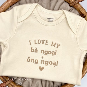 May include: A white baby onesie with the text "I love my bà ngoại + ông ngoại" embroidered in brown. A small brown heart is embroidered below the text.
