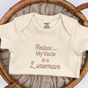 May include: A cream-colored baby onesie with the text "Relax... My Uncle is a Lineman" printed in brown. The onesie is laid out on a woven basket.