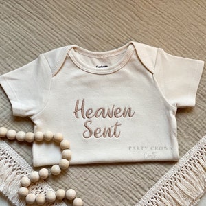 Puede incluir: Un body blanco para beb&eacute; con las palabras "Heaven Sent" bordadas en marr&oacute;n en la parte delantera. El body est&aacute; sobre una tela beige texturizada con un ribete de crochet blanco y una hebra de cuentas de madera.