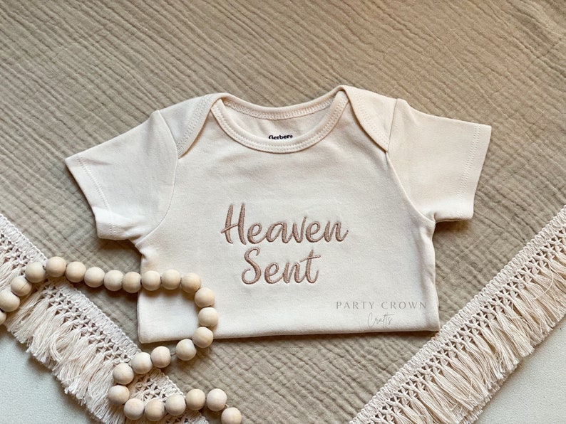Puede incluir: Un body blanco para beb&eacute; con las palabras "Heaven Sent" bordadas en marr&oacute;n en la parte delantera. El body est&aacute; sobre una tela beige texturizada con un ribete de crochet blanco y una hebra de cuentas de madera.
