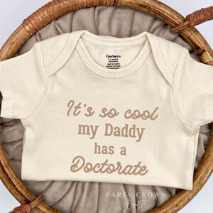 May include: Cream-colored baby onesie with the text "It's so cool my Daddy has a Doctorate" in brown lettering. The onesie is displayed in a woven basket with a neutral-colored fabric lining.