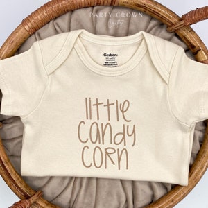 May include: A cream-colored baby onesie with the text "little candy corn" in brown lettering.