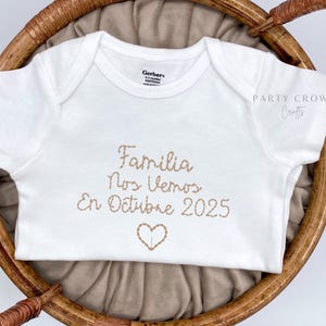 May include: White baby onesie with brown embroidered text that reads "Familia nos vemos en Octubre 2025" with a brown embroidered heart.