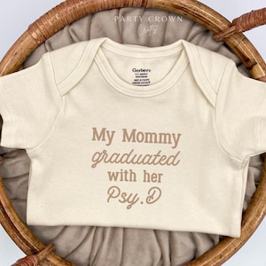 May include: Cream-colored baby onesie with the text "My Mommy graduated with her Psy.D" in brown lettering. The onesie is displayed in a woven basket. The Gerber brand label is visible.