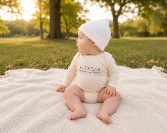 May include: A long-sleeved cream-colored baby bodysuit featuring a small black graphic of flowers and the text The Lord is my Shepherd, paired with a plain white ribbed knit beanie cap. The garment is made of soft fabric with a simple round neckline.