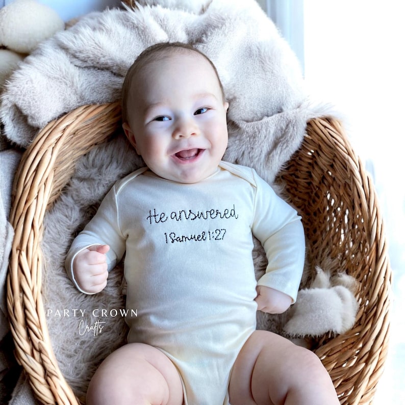 K&ouml;nnte beinhalten: Ein l&auml;chelndes Baby in einem cremefarbenen Body mit dem gestickten Text "He answered 1 Samuel 1:27". Das Baby liegt in einem Weidenkorb, der mit einer weichen, beigefarbenen Decke ausgekleidet ist. Die Worte "Party Crown Crafts" sind sichtbar.