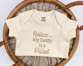Peut inclure: Un body bébé de couleur crème à manches courtes avec le texte "Relax... My Daddy is a Pilot" imprimé en brun.